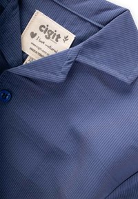 Zils svītrots audums ar apkakli, kurā ir apģērba etiķete ar uzrakstiem "cigit" un "Es mīlu dabu." Etiķete ir iestrādāta krekla audumā.