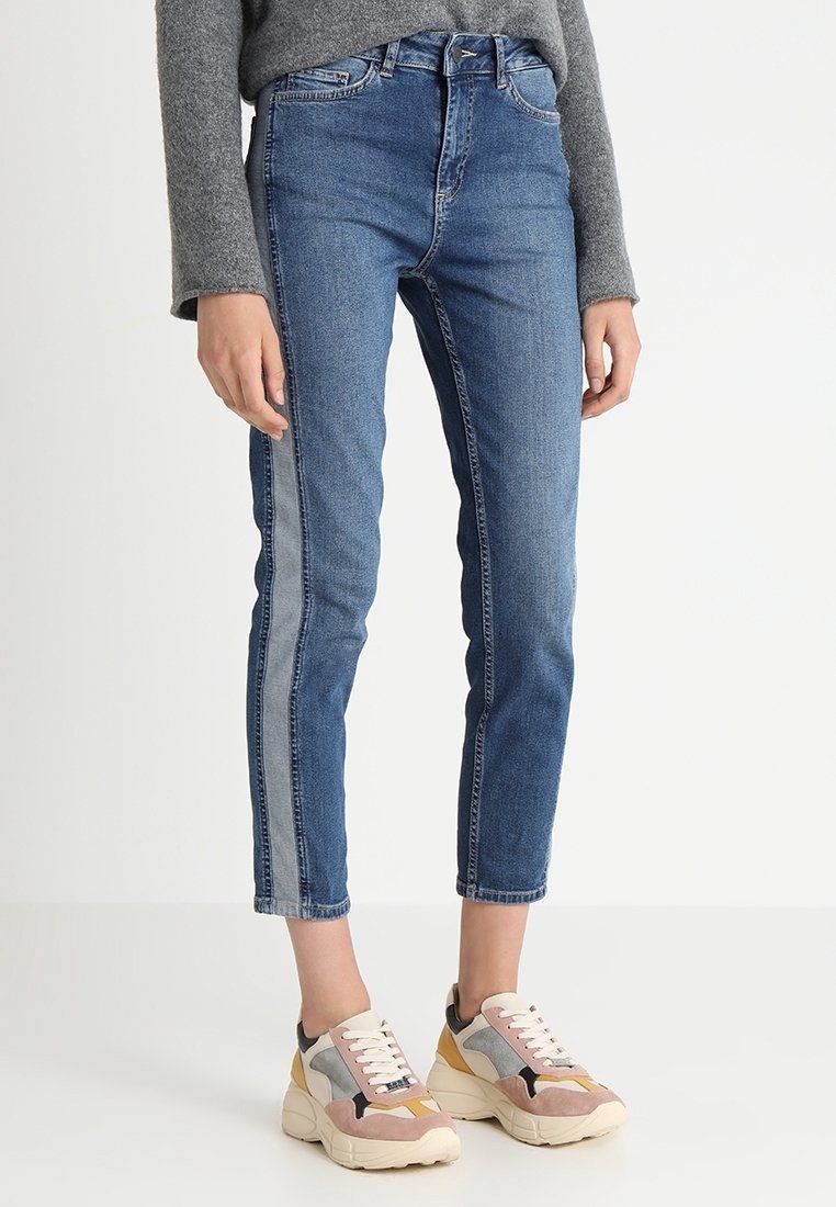 Personne portant un jean bleu avec une bande latérale bleu clair, associé à des baskets épaisses multicolores et un haut gris à manches longues.