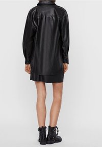 Robe chemise en cuir noir à coupe longue et décontractée, avec un dos boutonné et une texture lisse. Associée à des bottines noires avec des accents métalliques.