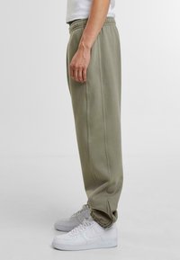 Pantaloni de trening de un verde măsliniu deschis, confecționați dintr-un material moale, având o croială relaxată, un talie elastică și detalii pe cusăturile laterale, purtați cu adidași albi.