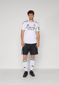 Biely futbalový dres s čiernymi a zlatými akcentmi, so značkou "Emirates", doplnený o čierne šortky a biele ponožky s čiernymi pruhmi.