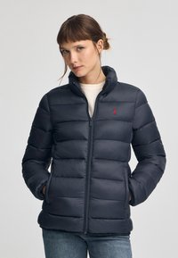 Marinblå pufferjacka med dragkedja, hög krage och ribbade muddar. Har sydda horisontella paneler och en liten röd logotyp på bröstet.