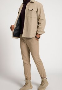 Beżowa kurtka koszulowa z mieszanki wełny z jedną kieszenią na piersi, czarnym guzikiem i miękką fakturą. W zestawie z dopasowanymi beżowymi spodniami i beżowymi tenisówkami.