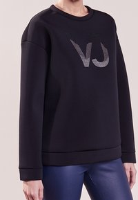 Sweatshirt noir avec un col rond, présentant un logo texturé en argent "VJ". Tissu doux, coutures d'épaule arrondies et poignets côtelés.