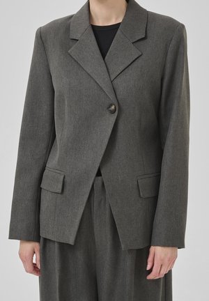 Femme portant une veste de costume grise ajustée avec un seul bouton et des poches à rabat, associée à un pantalon assorti et un haut noir.