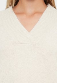 Maglione a V in morbido tessuto a maglia color crema; presenta un colletto a costine e una finitura testurizzata, evidenziando il suo design leggero e accogliente.