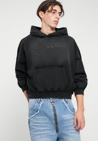 Črna oversized kapuca iz mehkega blaga z sprednjim žepom in embosiranim napisom. V kombinaciji z strganimi svetlo modrimi denim hlačami.