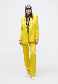 Žlté dvojradové sako a zodpovedajúce nohavice, doplnené čiernobielym vzorovaným topom a modrou vzorovanou čiapkou. Biele topánky.