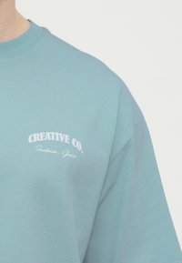 Κοντινό πλάνο ατόμου που φοράει ανοιχτό μπλε μπλουζάκι με άσπρο κείμενο "CREATIVE CO. Santorin_Greece" στο στήθος.