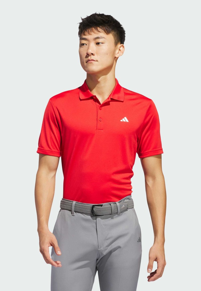 Rød polo skjorte med krave, korte ærmer og Adidas-logo. Sammen med lysegrå bukser og et gråt vævet bælte. Glat stoftekstur.