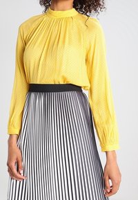 Blouse à manches longues à motifs jaunes avec un col haut ; associée à une jupe plissée à rayures noir et blanc avec une taille noire.