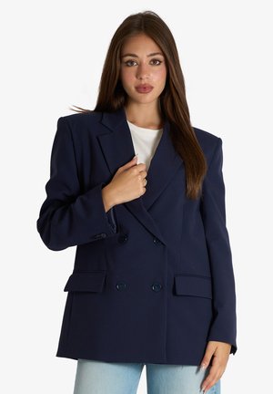Giovane donna con lunghi capelli castani che indossa un blazer doppiopetto blu navy oversize su una camicia bianca e jeans azzurri, che posa davanti a uno sfondo bianco.