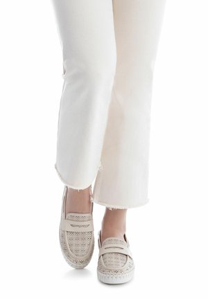Zapatos beige claro perforados tipo slip-on con suela plana blanca, combinados con jeans blancos de dobladillo deshilachado, mostrando un diseño relajado.
