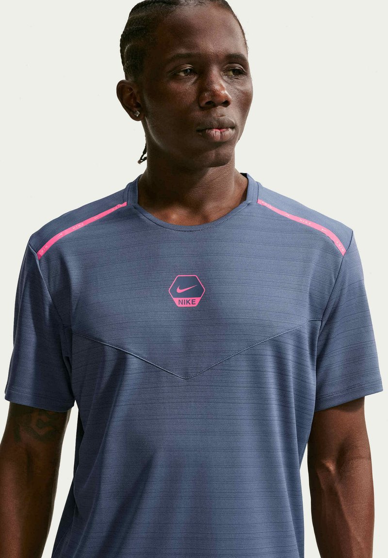 Hombre con trenzas que lleva una camiseta deportiva azul de Nike con detalles rosas, mirando hacia un lado sobre un fondo liso.