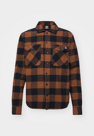 Chemise en flanelle à carreaux marron et noir avec une fermeture à boutons, deux poches poitrine et des manches longues ; présente un ourlet arrondi et des manches retroussées.