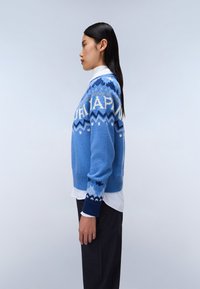 Maglione a maglia di colore blu chiaro con motivi geometrici, dotato di scollo rotondo e polsini a coste blu navy in contrasto. Indossato su una camicia bianca.