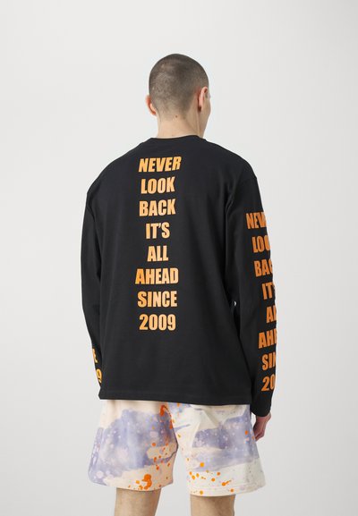 Chemise noire à manches longues avec texte orange au dos et sur les manches. Associée à un short de couleur claire avec des motifs éclaboussés de violet et d'orange.