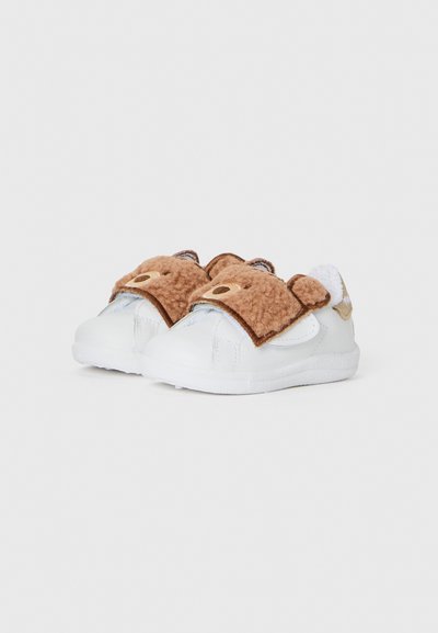 Des baskets blanches avec un visage de bear en peluche marron et des oreilles sur la sangle. Texture douce avec une semelle lisse et des accents en peluche.