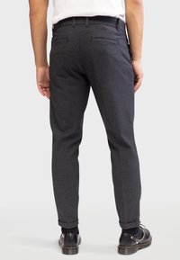Homme portant un pantalon gris foncé sur mesure avec des ourlets à revers, des chaussettes noires et des chaussures en cuir noires avec des détails en chaîne, vu de dos.