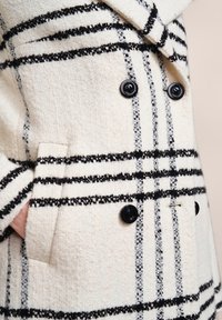 Doppelreihiger Wollmantel in Creme mit schwarz-grauem Karomuster, ausgestattet mit strukturiertem Stoff, zwei Fronttaschen und schwarzen Knöpfen.