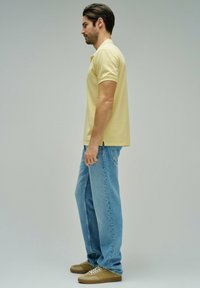 Hombre de perfil, vestido con una camiseta polo de manga corta color amarillo pálido, jeans azules y zapatillas marrones, sobre un fondo gris liso.