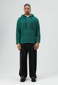 Sweat à capuche vert en tissu texturé et duveteux avec poche à l'avant. Associé à un jean noir ample et des chaussures noires avec des lacets roses.