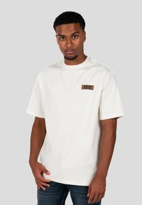 Kremowa koszulka z krótkim rękawem, wykonana z bawełny, z okrągłym dekoltem, posiada prostokątną brązową łatę z logo po lewej stronie klatki piersiowej.