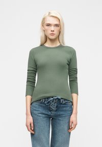 Ξανθιά γυναίκα με ριμπ πλεκτό πράσινο χακί μπλουζάκι με μακρύ μανίκι και μπλε τζιν με δαντέλα, στέκεται μπροστά σε λευκό απλό φόντο.