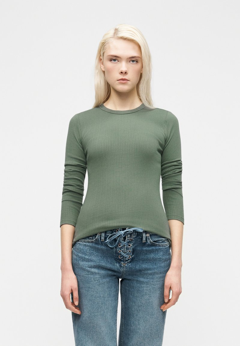 Ξανθιά γυναίκα με ριμπ πλεκτό πράσινο χακί μπλουζάκι με μακρύ μανίκι και μπλε τζιν με δαντέλα, στέκεται μπροστά σε λευκό απλό φόντο.