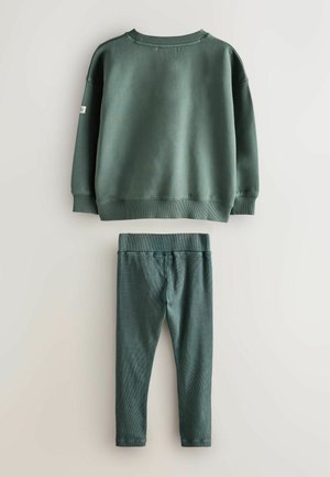 Tumma vihreä huppari, jossa on ribbireunat hihansuissa ja kauluksessa, yhdistettynä vastaaviin ribbireunaisiin legginsseihin. Pehmeä rakenne ja istuva muotoilu mukavuutta varten.