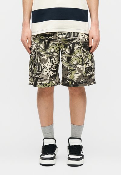 Άτομο που φοράει πράσινα, μαύρα και μπεζ καμουφλάζ cargo σορτς με τσέπη cargo, γκρι κάλτσες και ασπρόμαυρα αθλητικά παπούτσια.