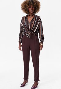 Vrouw met krullend haar, gekleed in een gestreepte bruine blouse die in de taille is gestrikt, bruine hoge taille broek en bordeauxrode puntige hakken, staand tegen een witte achtergrond.