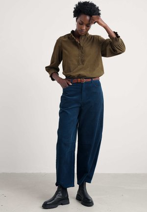 Olijfgroene blouse met ruffles en kraag, gecombineerd met donkerblauwe corduroy wijde broek en een gevlochten bruine riem, afgewerkt met zwarte enkellaarsjes.