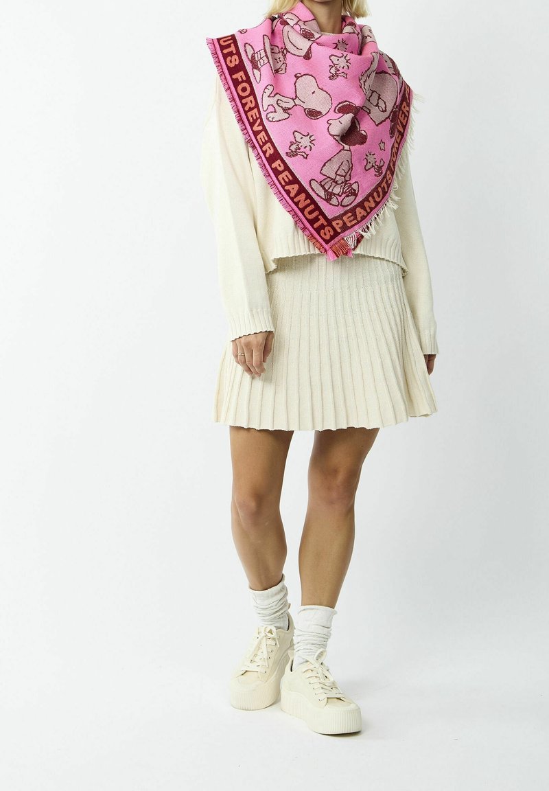 Femme portant une écharpe rose thème Peanuts avec Snoopy et Woodstock, un pull crème, une jupe plissée, des chaussettes blanches et des baskets crème, debout.