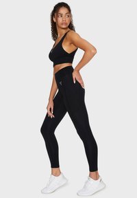 Mustat urheilutoppi paksuilla olkaimilla yhdistettynä korkeavyötäröisiin mustiin leggings-housuihin. Molemmissa on sileä pinta ja minimaalinen yksityiskohtaisuus. Valkoiset urheilukengät.