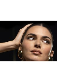 Close-up van een persoon met een egale huid, lichtbruine lipkleur, donkere eyeliner en grote gouden hoepeloorbellen. Het haar is strak naar achteren gekamd.