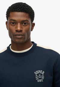 Sudadera azul marino con logo blanco y acentos en beige en los hombros. Presenta un cuello redondo y una tela texturizada.