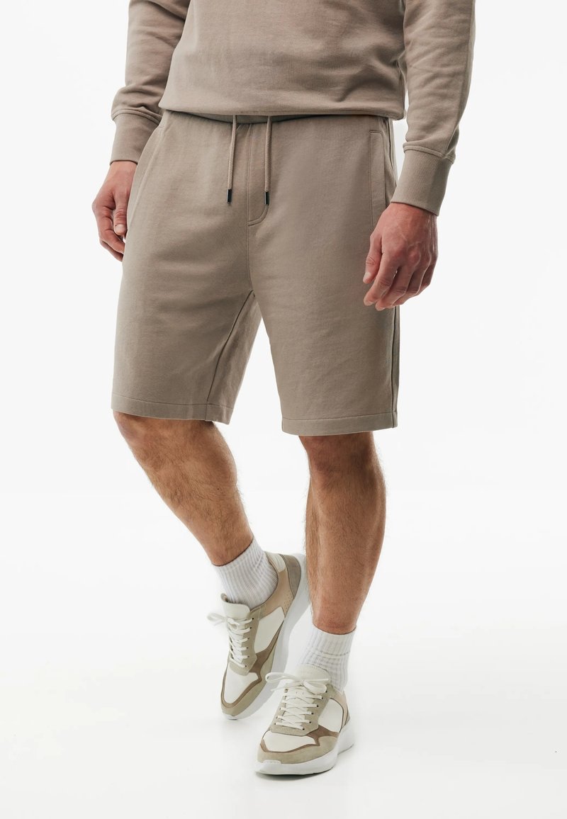 Man draagt beige shorts met trekkoord, bijpassende lange mouw top, witte sokken en beige-witte sneakers, staand op een witte achtergrond.