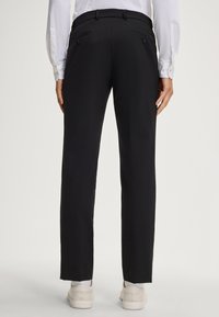 Zwarte broek met een getailleerde pasvorm, voorzien van twee achterzakken met knopen en een gladde textuur. G gedragen met een witte blouse met lange mouwen.