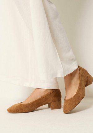 Füße tragen hellbraune Wildleder-Schuhe mit Blockabsatz, kombiniert mit locker sitzenden weißen Hosen vor einem neutralen Hintergrund.
