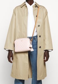 Person som bär en beige trenchcoat, en vit skjorta med knappar, blå jeans och bär en ljusrosa axelväska med fransdetaljer.
