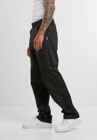 Black, loose-fitting pants with an elastic waistband, side pockets, and cuffed ankles, made of woven fabric. Worn with white sneakers.