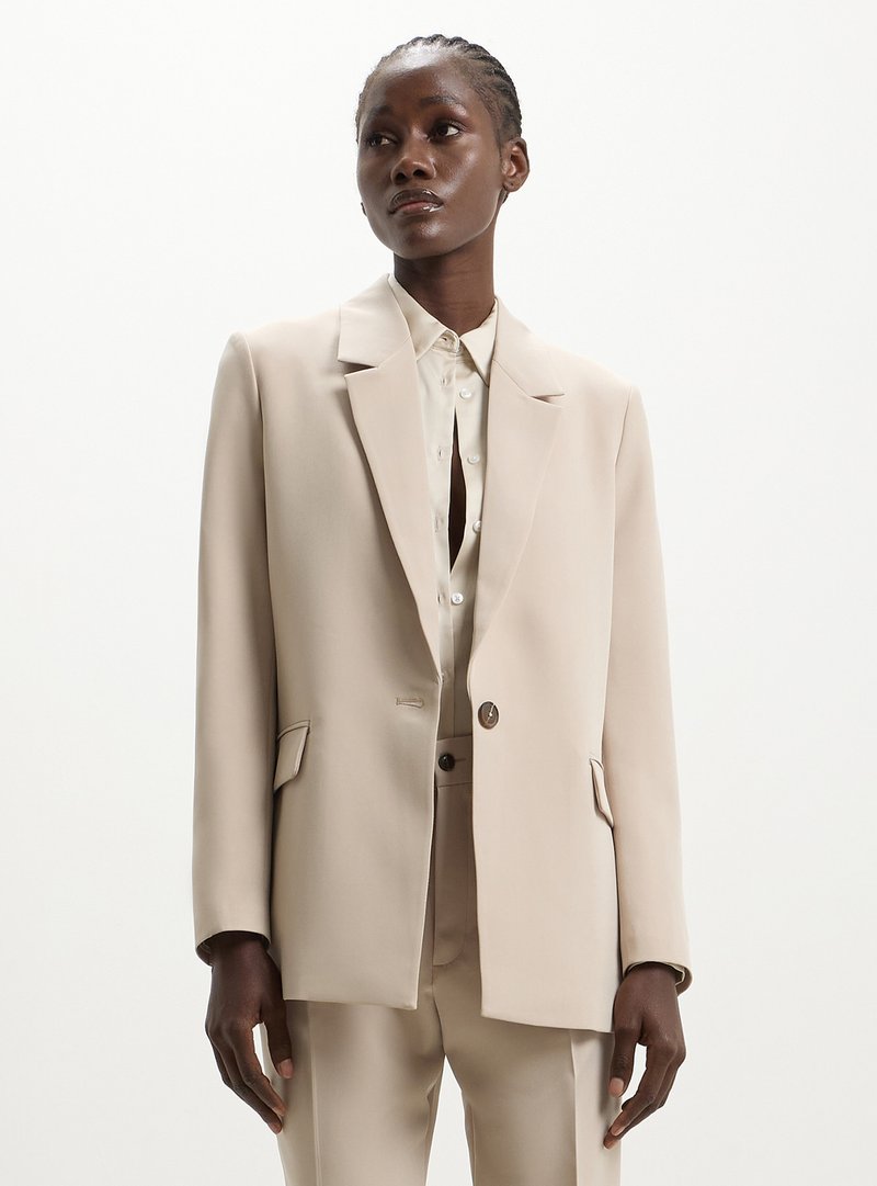 Blazer beige ajusté avec une silhouette structurée, des revers pointus, une fermeture à un bouton et des poches latérales, porté sur une chemise boutonnée claire.