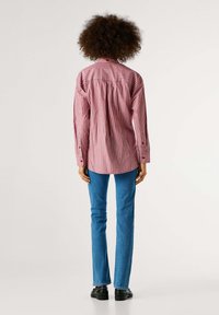 Camisa de rayas rojas y blancas con un corte relajado, con detalles en la yoke trasera, puños abotonados, combinada con jeans rectos azules y zapatos negros.