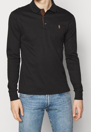Homme portant un polo noir à manches longues avec une patte de boutonnage marron et un jean bleu clair, debout devant un fond clair uni.