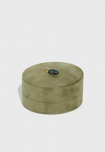 Okrogla kutija za nakit u maslinasto zelenoj zamši s glatkom teksturom, koja na vrhu ima mali tamnozeleni dragulj.