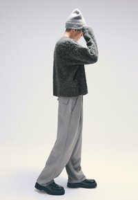 Sivý chlpatý sveter, sivé nohavice s širokými nohavicami a čierna obuv. Oblečenie obsahuje čiapku a voľný strih s textúrovanými materiálmi.