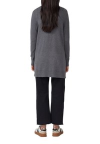 Personne avec de longs cheveux noirs portant un pull gris à manches longues, un pantalon noir à large jambe et des baskets blanches avec des semelles brunes, se tenant de dos.