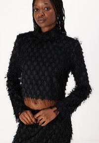Svart cropped topp med høy hals, med et teksturert mønster av hevede prikker og frynsete detaljer langs kantene og ermene.