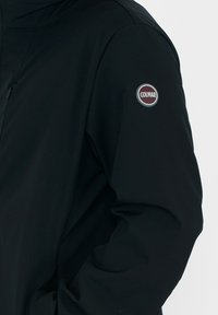 Chaqueta negra hecha de un tejido suave y resistente al agua. Presenta un bolsillo con cremallera y un logo circular en la manga. Diseño simple y funcional.